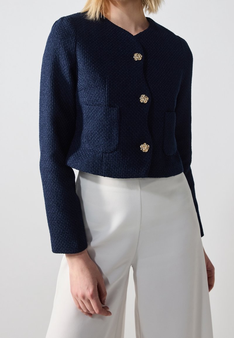 Femme portant un cardigan bleu marine texturé avec des boutons dorés et un pantalon blanc taille haute sur un fond uni.