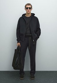 Jongeman in zwarte zonnebril, zwarte hoodie, zwarte broek en zwarte schoenen die een grote zwarte totebag tegen een effen muur houdt.