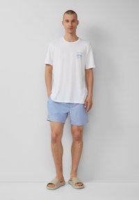 Weißes Baumwoll-T-Shirt mit blauem Grafikdruck auf der Brust, kombiniert mit hellblauen Shorts und beigen Slippern. Einfaches, lässiges Sommeroutfit.