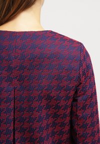 Textured blue fabric with a red geometric pattern, featuring short sleeves, a round neckline, and a central seam at the back.