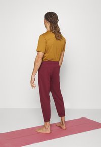 Bordeaux sweatpants met elastische boorden en een brede tailleband, gecombineerd met een T-shirt met korte mouwen in mosterdkleur. Blote voeten op een roze yogamat.