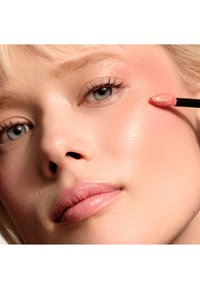 Gros plan sur un visage appliquant un produit rose brillant sur la joue à l'aide d'un applicateur noir. La peau a une texture lisse et déwy.