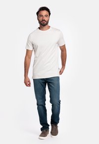 Weißes Kurzarm-T-Shirt mit Rundhalsausschnitt, kombiniert mit dunkelblauen Jeans. Das Modell trägt graue Sneakers und steht vor einem schlichten Hintergrund.
