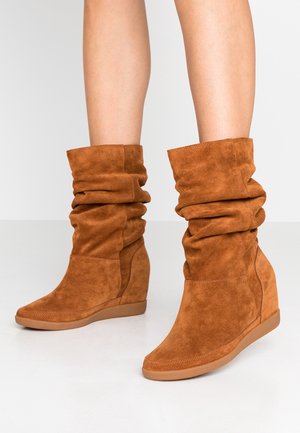 Botines de cuña con tacón ancho de ante marrón, de aspecto holgado, usados sobre piernas desnudas, con suelas de goma texturizadas sobre un fondo claro.