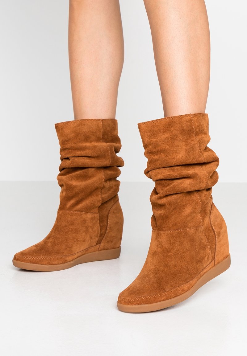 Bottes compensées marron en daim souple portées sur jambes nues, avec semelles en caoutchouc texturées sur un fond clair.