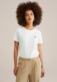 Balta medvilnės t-shirt su trumpomis rankovėmis, su mažuoju siuvinėtų logotipu diamanto forma ant krūtinės. Derinama su šviesiai rudomis kelnėmis.