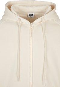 Sudadera con cremallera color crema, hecha de una tela suave, con capucha ajustable y detalles acanalados a lo largo de las costuras. Etiqueta visible en el interior.