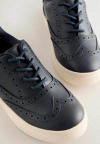 Tumši zila ādas sporta apavi ar brogue detaļām, atbilstošām tumši zilām šņorēm un krēmkrāsas gumijas zolīti. Gluda tekstūra ar perforētām detaļām.