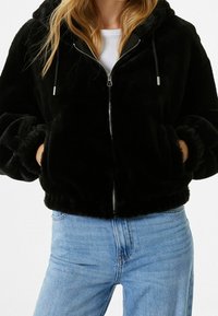 Svart fuskpälsjacka med dragkedja och huva, med ribbade ärmar, sidofickor och mjuk textur, i kombination med ljusblå jeans.