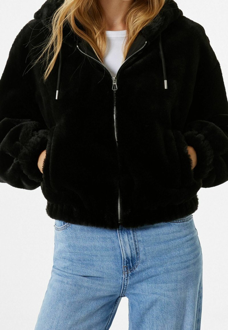 Svart fuskpälsjacka med dragkedja och huva, med ribbade ärmar, sidofickor och mjuk textur, i kombination med ljusblå jeans.