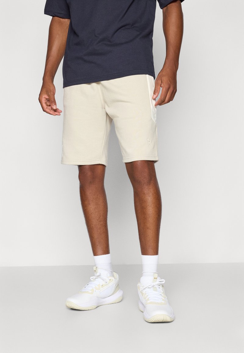 Beige treningshortser laget av mykt stoff med sidelommer, kombinert med en mørk blå t-skjorte og hvite sko med gule detaljer.