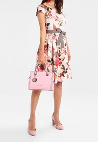 Donna che indossa un vestito floreale con un fiocco in vita grigio, tiene una borsa rosa e indossa dei tacchi alti rosa abbinati, in piedi su uno sfondo bianco.