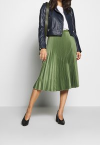 Giacca da biker in pelle blu navy, t-shirt bianca, gonna midi plissettata verde e tacchi neri testurizzati. La giacca presenta cerniere e un design aderente.