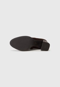 Vue de la semelle d'une chaussure en cuir marron foncé avec une semelle adhérente texturée et un talon bloc sur un fond clair uni.