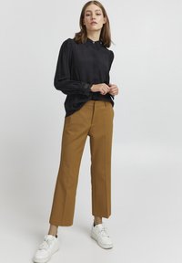 Blouse noire à manches longues avec une finition texturée et des détails en dentelle au niveau du collier et des poignets, associée à un pantalon tailleur couleur moutarde et des baskets blanches.