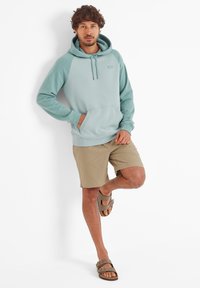Man in teal hoodie, beige shorts, and sandals leaning against a white wall with one leg bent, hands in pocket and on thigh.