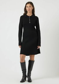 Abito a maglia a coste nero con colletto, placca di bottoni e maniche lunghe. Indossato con stivali di pelle nera al ginocchio. Orlo dritto.