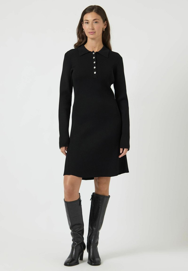 Abito a maglia a coste nero con colletto, placca di bottoni e maniche lunghe. Indossato con stivali di pelle nera al ginocchio. Orlo dritto.