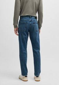 Dark blue denim jeans with a straight-leg cut, featuring two back pockets and visible stitching. Worn with a gray long-sleeve top and white shoes.
