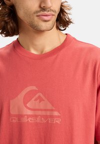 Korálové tričko vyrobené z měkké bavlny, které má na přední straně velké, světlé logo Quiksilver a grafiku hor. Kolo krku.