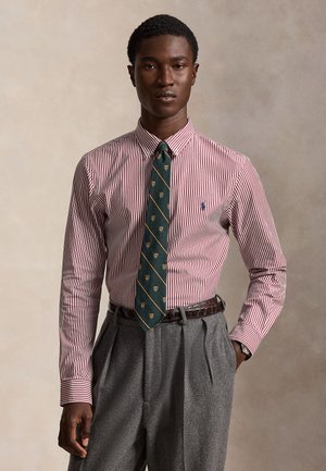 Jeune homme portant une chemise rayée rouge et blanche avec une cravate verte à motifs et un pantalon gris plissé, debout devant un fond beige.