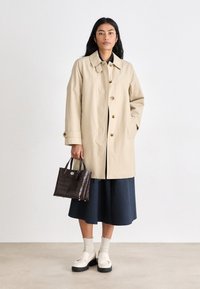 Beige trench coat with large buttons, navy skirt, cream socks, and black platform shoes. Holding a dark brown croc-embossed handbag.
