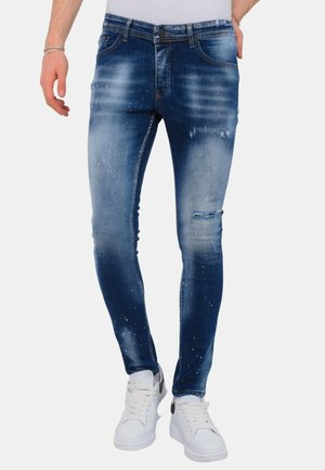 Slim-fit blauwe jeans met vervaagde en versleten details, gecombineerd met witte sneakers en een wit topje, gedragen door een staande persoon.