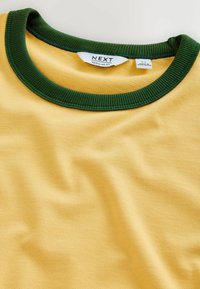 T-shirt jaune avec col rond côtelé vert foncé et étiquettes visibles « NEXT Regular Fit » et pays de fabrication.