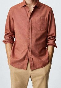 Raskahvi värvi nupp-särk, millel on tasku ja pika käe rulli, valmistatud tekstuurist kangast. Kanda beezide puuvillaste voodipükste peal.