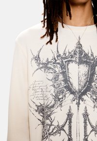 Άτομο που φοράει μια κρεμ πουκαμίσα με υφή και σχέδιο μαύρης αγκαθωτής ασπίδας, καθώς και ασημένιο κολιέ με μενταγιόν στο σύμβολο του Ματιού του Ώρου.