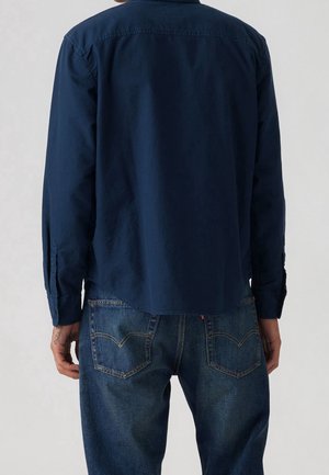 Mies, joka pukeutuu tummansiniseen pitkähihaiseen paitaan ja sinisiin farkkuihin, kuvattu takaa yksinkertaisen taustan edessä.