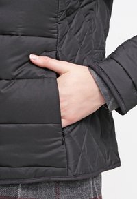 Main insérée dans la poche latérale d'une veste matelassée noire avec coutures en losanges et détail zippé.