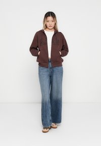 Brown zip-up hoodie with "GAP" in black print, featuring a hood and front pockets, paired with wide-leg faded blue jeans.