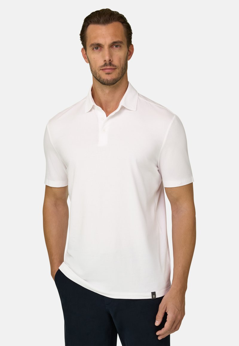 Homme aux cheveux bruns courts et à la barbe, portant un polo blanc uni à manches courtes et un pantalon foncé, debout avec une main dans la poche.
