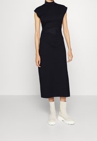 Černé, přiléhavé midi šaty s krátkými rukávky a stojáčkem. Mají texturovaný černý pás. Nosené s krémovými kotníkovými botami.