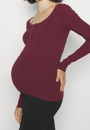 Zwangere persoon in een donkerrood shirt met lange mouwen en een zwarte rok, hand rustend op de onderrug, torso en buik zichtbaar, neutrale achtergrond.