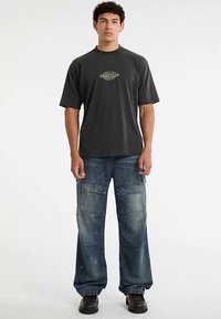 Graues T-Shirt mit kurzen Ärmeln und "ORIGINALS"-Grafik sowie locker sitzende blaue Jeans-Cargohosen mit mehreren Taschen. Schwarze Schuhe.