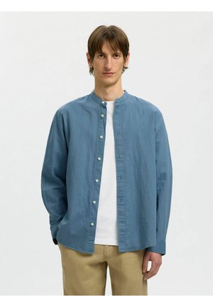 Junger Mann mit glattem braunem Haar, der ein blaues Hemd über einem weißen T-Shirt und beige Hosen trägt, vor einem weißen Hintergrund stehend.