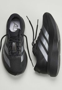 Ζευγάρι μαύρα και γκρι αθλητικά παπούτσια Adidas με το ένα παπούτσι δεμένο και το άλλο λυμένο σε ανοιχτό φόντο.