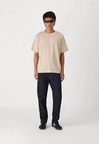 Bēša kokvilnas t-krekls ar īsām piedurknēm, kombinēts ar tumši zilām džinsa biksēm un melniem sporta apaviem ar baltām svītrām. Modelis valkā melnas saulesbrilles.