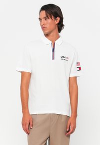 Hvid polo skjorte med krave, marineblå detaljer på placket, broderet "USA" logo og flaglapper på ærmerne. Korte ærmer.