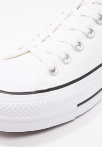 Sneaker bianca in tela con punta in gomma, occhielli argentati, lacci bianchi, suola bianca texturizzata e una sottile striscia nera lungo il fondo.