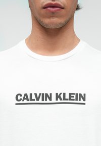 Bela majica s kratkimi rokavi, ki se ponaša z odširnim črnim napisom "CALVIN KLEIN" čez prsni del, okroglim izrezom in mehkobo bombažne tkanine.