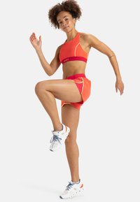 Sportbeha en shorts in feloranje met rode accenten. De beha heeft een logo, terwijl de shorts een losse pasvorm hebben. Witte sportschoenen maken de outfit compleet.