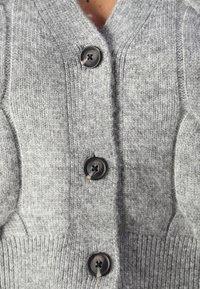 Grijze gebreide cardigan met een V-hals, voorzien van vier zwarte knopen, geribde zoom en een gestructureerde stof. Geen zichtbare patronen of accenten.