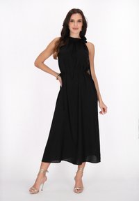 Schwarzes ärmelloses Kleid mit hohem Ausschnitt, gerafften Details und seitlichen Bindebändern. Kombiniert mit metallischen Riemchenschuhen. Glattes Material.