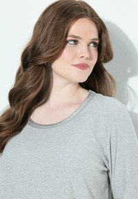 Frau mit langen braunen Haaren, die ein hellgraues, geripptes Langarmshirt trägt und leicht nach rechts schaut, vor einem weißen Hintergrund.