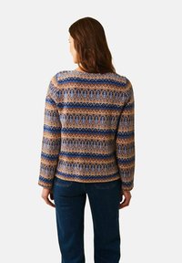 Kvinna med långt brunt hår, iklädd en blå, brun och beige mönstrad tröja och mörkblå jeans, stående med ryggen mot betraktaren på vit bakgrund.