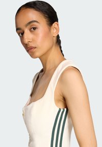 Svetlý béžový šaty s obdĺžnikovým výstrihom, ramienkami a zelenými vertikálnymi pruhmi po stranách. Obsahuje detail zlatého gombíka.