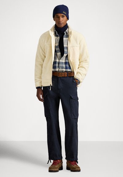 Άνδρας που φοράει μπεζ φλις μπουφάν, μπλε καρό πουκάμισο, ναυτικό παντελόνι cargo, καφέ ζώνη, μπλε σκουφάκι και καφέ μποτάκια πεζοπορίας με κόκκινες κορδόνια που στέκεται.